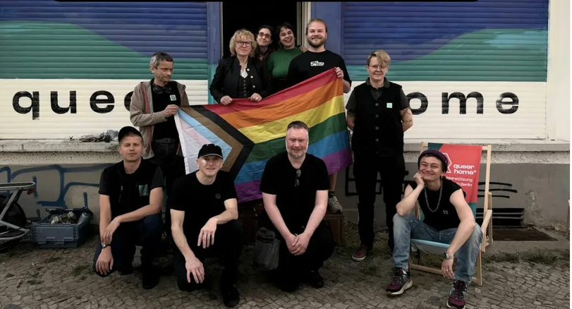 Team von queerhome* mit Progress-Pride-Flagge vor dem queerhome*-Standort in Berlin. Zu Besuch ist Sabine Pöhl, Diversity- und Queer-Beauftragte des Bezirks Lichtenberg, anlässlich des Internationalen Tags gegen Queerfeindlichkeit am 17. Mai 2026.